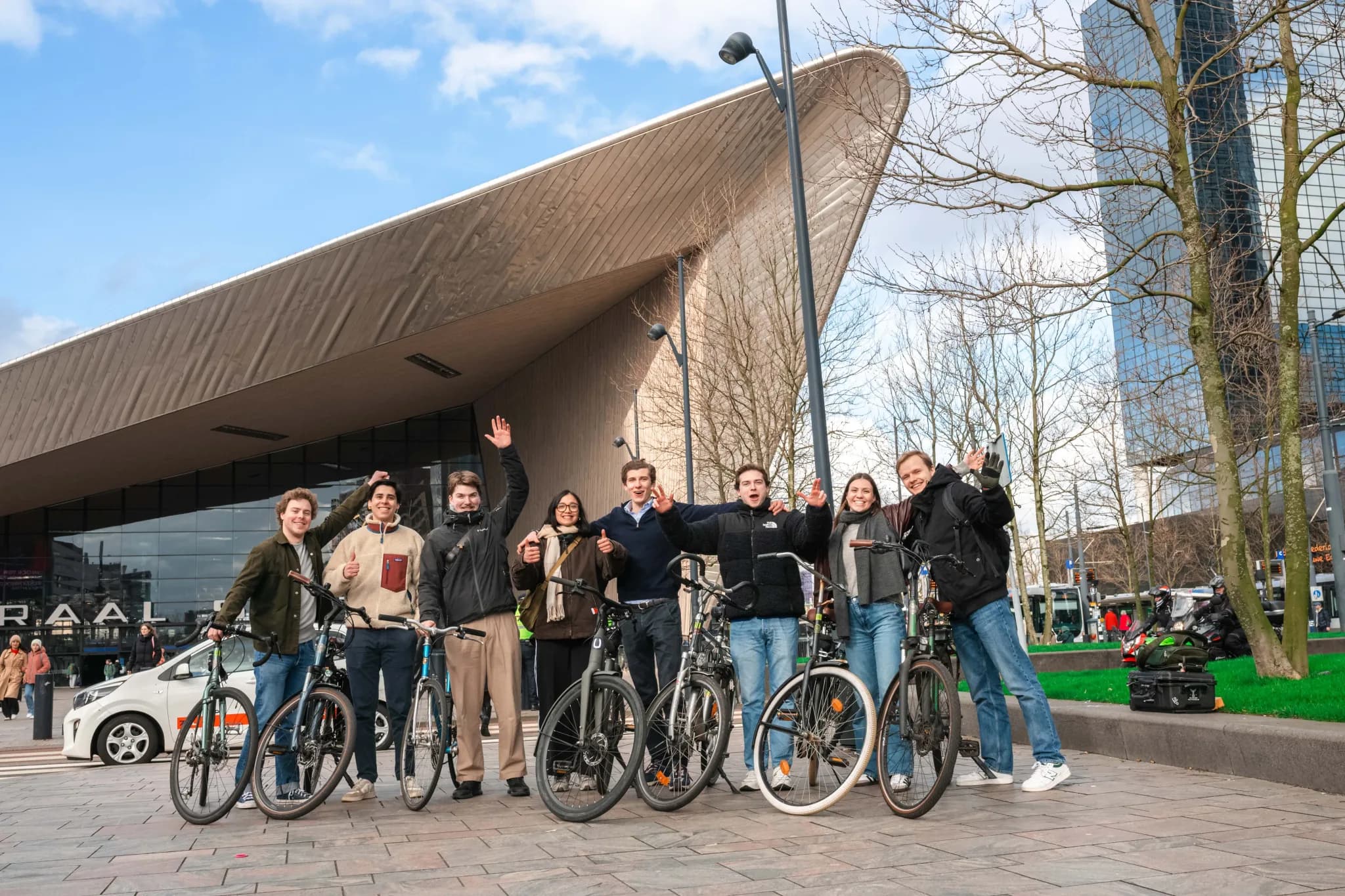 BIKEBAZE team members waving with their bikes in front of the curved roof of Rotterdam Central Station.