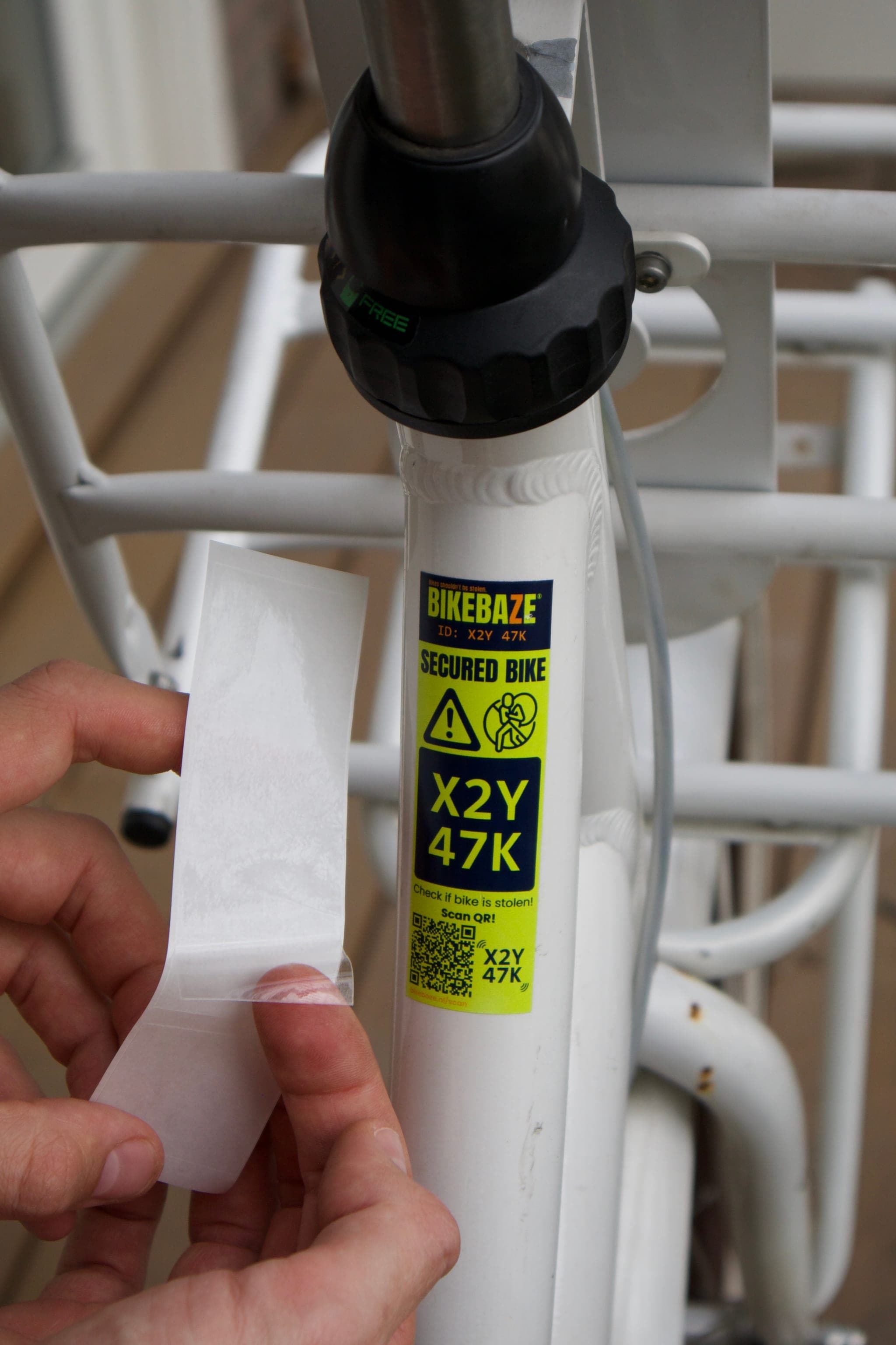 Hand applying a clear SafeTag Protect overlay onto a yellow BIKEBAZE SafeTag on a white bike frame.